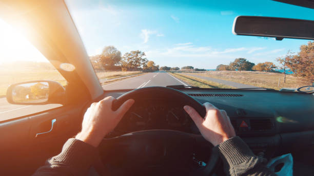 Personal perspective - point of view (POV) - of driver, who drives his car on a road. The hands are on the steering wheel and the sun is shining outside. He drives on country roads as the sun is about to set. The sky is blue.