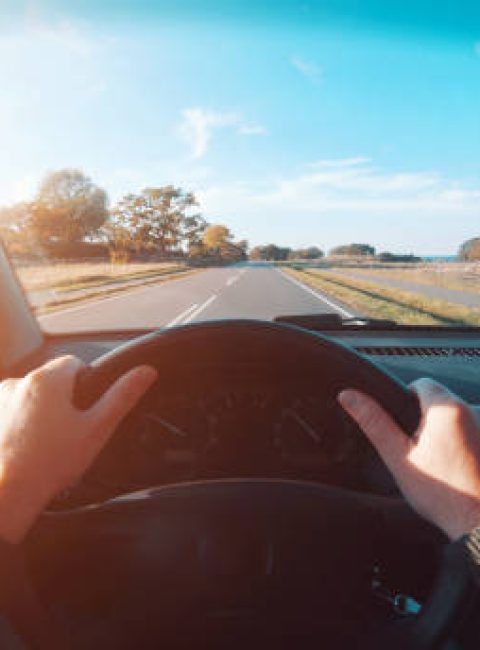 Personal perspective - point of view (POV) - of driver, who drives his car on a road. The hands are on the steering wheel and the sun is shining outside. He drives on country roads as the sun is about to set. The sky is blue.